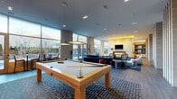 This single-floor amenity lounge is a well-appointed communal space designed with a contemporary aesthetic and a bold, curated interior style. The open layout is divided into distinct functional zones that flow seamlessly into one another, creating a cohesive and inviting atmosphere throughout.

The main lounge area anchors the space with a generous sectional sofa in deep navy blue, accented by colorful throw pillows and arranged atop a striking large-format area rug with an abstract design. A fireplace and wall-mounted television are framed by a backlit built-in shelving unit finished in warm yellow tones, adding a vibrant focal point to the room. Decorative vertical column accents in textured stone further define the space and add architectural interest.

A billiards table occupies its own dedicated zone, illuminated by modern pendant lights and bordered by patterned carpet. Adjacent to this is a dining and kitchenette area featuring hexagonal tile flooring, a long communal table, pendant lighting, and a full cabinetry run with a wet bar, beverage refrigerator, and a reflective tile backsplash.

Floor-to-ceiling glass walls and French doors flood the interior with natural light and provide direct access to an outdoor patio, where views of a pool area and pergola structure are visible. The overall ambiance is energetic yet comfortable, blending bold design choices with practical amenities suited for social gatherings.