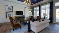 This thoughtfully designed single-floor space makes a striking first impression with its sophisticated living area. The focal point of the room is a dramatic navy blue tray ceiling, which adds depth and a bold design statement, complemented by an elegant rustic chandelier that casts warm ambient light throughout the space. Large windows dressed with dark curtain panels frame views of the surrounding exterior, flooding the interior with generous natural light and creating an airy, open atmosphere. The living area is furnished with a classic white tufted sofa, rich brown leather wingback chairs, and woven accent seating, offering a layered and inviting aesthetic. A wall-mounted flat-screen television anchors one side of the room above a low-profile media console, while a rustic wooden coffee table grounds the seating arrangement. A sliding barn door adds a charming architectural detail and a nod to contemporary farmhouse style. Wood-look flooring runs throughout, providing warmth underfoot, while a blue area rug ties the color palette together cohesively. The overall ambiance blends coastal, traditional, and modern elements into a welcoming and well-appointed living environment.