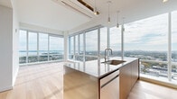 Located at 50 Clinton Street in New Rochelle, NY, this modern 1-bedroom, 2-bathroom residence offers an elevated living experience defined by sleek design and breathtaking views. Light hardwood flooring flows throughout the open-plan layout, complemented by crisp white walls and abundant natural light pouring in through dramatic floor-to-ceiling windows. The expansive living areas provide sweeping panoramic views of the surrounding cityscape, Long Island Sound, and distant Manhattan skyline, creating a striking backdrop from nearly every room. The contemporary kitchen is outfitted with warm wood cabinetry, stainless steel countertops and appliances — including a high-end refrigerator and gas range — and a central island with an undermount sink, all framed by more spectacular views. The well-proportioned primary bedroom enjoys generous natural light through large windows overlooking the city below. Two thoughtfully designed bathrooms feature large-format tile flooring, wood vanities with white countertops, illuminated mirrors, and clean, modern fixtures — one with a soaking tub and one with a glass-enclosed walk-in shower. A dedicated closet and multiple living areas round out this refined and functional high-rise residence.