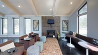 This single-story space is designed with an open, airy layout that makes a strong architectural impression. Dramatic floor-to-ceiling windows flood the interior with natural light and offer views of surrounding greenery and the neighborhood streetscape, creating a seamless connection between indoors and outdoors. The soaring vaulted ceilings, accented by exposed beams and ceiling fans, amplify the sense of openness throughout.

Dark hardwood flooring runs continuously across the space, grounding the bright, neutral palette and adding warmth. A welcoming lounge area features brown leather seating arranged around low round tables, with patterned accent pillows providing a pop of texture. Adjacent to this is a fireplace framed by a full wood-panel surround, flanked by artwork and complemented by a plush area rug and eclectic ottomans — making it an inviting focal point for gathering.

The dining area showcases chartreuse accent walls that energize the space, with modern round pedestal tables paired with sleek white chairs and bar-height seating along a window-side counter. Pendant lighting adds a refined touch overhead. A glimpse into an adjoining kitchenette area suggests additional functionality, making this a well-rounded, versatile communal environment.