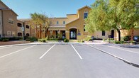 This single-story clubhouse serves as the central amenity hub of a Mediterranean-inspired residential community. The exterior showcases a warm golden stucco façade, a clay tile roof, arched entryways, and lush landscaping with mature trees, creating an inviting curb appeal complemented by ample parking.

Inside, the main lounge area is generously proportioned and designed for comfort and social gathering. The space features dual ceiling fans, recessed skylight-style panel lighting set within a tray ceiling, and wall-mounted televisions for entertainment. Two tufted, high-back sofas face each other across a dark wood coffee table, anchored by a patterned area rug. Natural light filters through multiple windows and a set of glass-paned double doors that open to an outdoor balcony view. A shelving display unit occupies one corner, adding a decorative touch.

The outdoor amenity area is a standout feature, centered around a large, freeform swimming pool with crystal-clear blue water. Surrounding the pool is a spacious deck furnished with lounge chairs, tables, and umbrellas, all framed by tall swaying palm trees. The multi-story residential buildings encircle the courtyard, offering a resort-like atmosphere ideal for relaxation and community interaction.