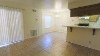 This single-floor residence offers a straightforward and functional layout designed for comfortable everyday living. The open dining and living area serves as the heart of the home, featuring large neutral-toned tile flooring that extends throughout the space, lending a clean and cohesive look. A central window allows natural light to brighten the room, enhancing the airy feel of the white walls. An elegant chandelier hangs overhead, adding a touch of classic charm to the dining area. The space transitions seamlessly into the kitchen via a raised counter with bar-style overhang, ideal for casual dining or food preparation. Dark cabinetry visible in the kitchen provides a subtle contrast to the otherwise light and neutral palette of the home. Climate control is well-integrated, with visible HVAC vents and a wall-mounted heating unit ensuring year-round comfort. The clean lines, open flow, and practical features of this single-story home make it a welcoming and versatile living space suited for a variety of lifestyles.