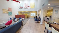This single-floor space presents a spacious, open-plan lobby with an airy, high-ceiling design that immediately conveys a sense of openness and warmth. Large light-toned tile flooring runs throughout the main area, reflecting the abundant ambient light and contributing to the clean, modern aesthetic.

The seating areas are thoughtfully arranged to accommodate a variety of needs. A casual lounge zone features a navy blue sofa paired with a wooden coffee table and a neutral area rug, while a semi-private booth with rich red upholstered banquette seating offers a more intimate setting. For those seeking a more structured setting, round tables with bold blue chairs provide a casual dining or meeting option.

Decorative elements elevate the space with personality and visual interest. A set of three vibrant canvas artworks — featuring teal, red, and yellow tones — adorn the upper walls, complemented by striking red origami-style bird sculptures suspended from the ceiling, adding a dynamic, artistic focal point overhead.

At the rear of the lobby, a reception or service counter is visible beneath warm yellow accent walls, creating a welcoming focal point for visitors. A small refreshment or service station is accessible along one side wall, adding to the functional convenience of the space. Overall, the design balances practicality with a lively, cheerful ambiance.
