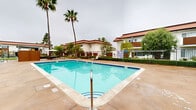 Located at 201 Los Arbolitos Boulevard in Oceanside, California, this single-level outdoor amenity space is the social heart of a well-maintained residential community. The centerpiece of the area is a generously sized swimming pool with clear, blue-green water, framed by a spacious concrete deck that provides ample room for lounge chairs and outdoor furniture. Adjacent to the pool sits a tiled spa, offering a dedicated space for relaxation and hydrotherapy. The pool area is enclosed by a wrought iron fence, ensuring safety while maintaining an open, airy feel. A separate seating area with a table and chairs provides a shaded spot for outdoor dining or socializing. The surrounding landscape is thoughtfully maintained, featuring tall, iconic palm trees, manicured shrubs, and lush green lawns that add to the Southern California aesthetic. A children's playground is visible in the background, making this a family-friendly environment. The residential buildings surrounding the courtyard feature a classic Spanish-inspired architectural style, with white stucco exteriors and terracotta tile rooflines that complement the warm, coastal ambiance of the Oceanside setting.