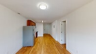 This single-floor residence offers a clean, functional layout unified by warm honey-toned hardwood flooring that extends throughout the space. The kitchen is well-appointed, featuring rich medium-toned wood cabinetry spanning both upper and lower sections, light countertops with a double-basin stainless steel sink, and a full suite of stainless steel appliances including a refrigerator, electric range, and over-the-range microwave. A long, open corridor connects the kitchen to the entry door, with a visible electrical panel mounted along the wall and a linear overhead light fixture providing bright, even illumination.

The bedroom is a comfortably sized, light-filled room with a window allowing natural light, white walls, and a set of double bifold closet doors offering generous storage space. The room connects directly to the kitchen area, creating a practical flow throughout the unit.

The bathroom features a full tub and shower combination enclosed by clean white subway-style tiles, with built-in corner shelving and a recessed soap dish for added functionality. White walls and crisp finishes give the bathroom a fresh, minimal appearance. Throughout the unit, the consistent use of white walls, white trim, and warm flooring creates a cohesive and inviting atmosphere.