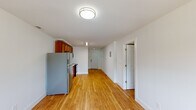 This single-floor residence offers a clean, functional layout unified by warm honey-toned hardwood flooring that extends throughout the space. The kitchen is well-appointed, featuring rich medium-toned wood cabinetry spanning both upper and lower sections, light countertops with a double-basin stainless steel sink, and a full suite of stainless steel appliances including a refrigerator, electric range, and over-the-range microwave. A long, open corridor connects the kitchen to the entry door, with a visible electrical panel mounted along the wall and a linear overhead light fixture providing bright, even illumination.

The bedroom is a comfortably sized, light-filled room with a window allowing natural light, white walls, and a set of double bifold closet doors offering generous storage space. The room connects directly to the kitchen area, creating a practical flow throughout the unit.

The bathroom features a full tub and shower combination enclosed by clean white subway-style tiles, with built-in corner shelving and a recessed soap dish for added functionality. White walls and crisp finishes give the bathroom a fresh, minimal appearance. Throughout the unit, the consistent use of white walls, white trim, and warm flooring creates a cohesive and inviting atmosphere.