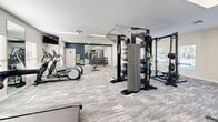 Located at 3325 Oak Forest Dr in Fayetteville, NC, this fitness center offers a comprehensive and thoughtfully organized workout facility spread across a single floor. The space is designed to accommodate a wide range of fitness activities, from cardiovascular training to strength and functional fitness.

The cardio zone features a row of treadmills and spinning bikes positioned along the windowed wall, where natural light filters in through multiple windows fitted with blinds. Wall-mounted televisions provide entertainment during workouts. Large mirrors are strategically placed throughout to enhance the sense of space and allow for form monitoring.

The strength training area is well-appointed with a multi-functional cable and pulley machine system, a dedicated free weight section with a full rack of dumbbells, stability balls, resistance bands, and a weight bench with additional attachments. The functional fitness zone includes an air bike, step platforms, and battle ropes for high-intensity training.

The flooring throughout consists of durable, patterned carpet tiles in a neutral gray tone, complementing the light-colored walls with a navy blue accent wall near the free weight area. Recessed panel lighting ensures the space is well-lit throughout. The open floor plan allows for easy navigation and simultaneous use of multiple equipment stations, making this fitness center a versatile and fully equipped facility.