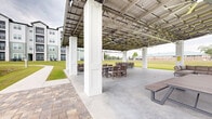 Located in Santa Rosa Beach, Florida, this community amenity space is a generously sized covered outdoor pavilion designed for gathering, relaxation, and recreation. The structure features a metal roof supported by tall white columns, creating an open-air environment that remains sheltered from the elements. Multiple ceiling fans are mounted throughout, ensuring comfortable airflow during warm Florida days, while string lights add ambiance for evening use. The concrete floor accommodates a variety of seating arrangements, including cushioned sectional sofas, lounge chairs, and picnic-style dining tables with benches — offering ample space for both casual socializing and larger gatherings. The pavilion opens on all sides to well-maintained green lawns and natural Florida landscape, with tall pines framing the horizon. A paver walkway connects the pavilion to a charming wooden dock that extends over a serene pond, complete with an active fountain feature that enhances the tranquil waterfront setting. Views from the pavilion capture the surrounding residential buildings and open green fields. The combination of shaded comfort, open sightlines, water views, and thoughtfully arranged outdoor furnishings makes this pavilion a standout community feature in the Santa Rosa Beach area.