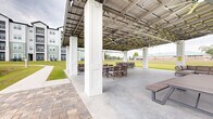 Located in Santa Rosa Beach, Florida, this community amenity space is a generously sized covered outdoor pavilion designed for gathering, relaxation, and recreation. The structure features a metal roof supported by tall white columns, creating an open-air environment that remains sheltered from the elements. Multiple ceiling fans are mounted throughout, ensuring comfortable airflow during warm Florida days, while string lights add ambiance for evening use. The concrete floor accommodates a variety of seating arrangements, including cushioned sectional sofas, lounge chairs, and picnic-style dining tables with benches — offering ample space for both casual socializing and larger gatherings. The pavilion opens on all sides to well-maintained green lawns and natural Florida landscape, with tall pines framing the horizon. A paver walkway connects the pavilion to a charming wooden dock that extends over a serene pond, complete with an active fountain feature that enhances the tranquil waterfront setting. Views from the pavilion capture the surrounding residential buildings and open green fields. The combination of shaded comfort, open sightlines, water views, and thoughtfully arranged outdoor furnishings makes this pavilion a standout community feature in the Santa Rosa Beach area.