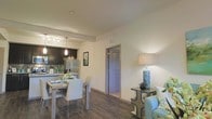 This thoughtfully designed single-floor residence showcases an open-concept layout that seamlessly connects the living, dining, and kitchen areas, creating a spacious and cohesive living environment. The kitchen is outfitted with dark cabinetry, a striking mosaic tile backsplash, stainless steel appliances including a French door refrigerator, and pendant lighting that adds a modern touch. A breakfast bar provides additional seating and separates the kitchen from the dining area, which features a light wood dining table with white chairs — a clean, contemporary aesthetic. The living area is warm and welcoming, furnished with a light blue accent chair and complementary side table, anchored by a decorative lamp that contributes to the cozy atmosphere. Throughout the space, rich wood-look hardwood flooring ties the rooms together and adds warmth underfoot. The neutral, light-toned walls enhance the sense of openness and allow natural and artificial light to reflect evenly. A hallway leads to additional rooms beyond, suggesting a practical and functional floor plan. Overall, this space strikes a balance between modern design and everyday comfort.