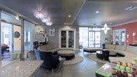 This sophisticated single-floor lounge space makes an immediate impression with its thoughtfully curated interior design and upscale finishes. The open-plan layout is anchored by two striking crystal chandelier fixtures that cascade elegantly from a tray ceiling, casting a warm glow throughout the room. Recessed lighting complements the chandeliers, ensuring the space is well-lit at all times.

The seating area features a curved sectional sofa paired with a round ottoman and woven accent chairs, all arranged atop circular area rugs that define the lounge zones. A second seating grouping with a low ottoman and dark accent chairs offers an additional area for gathering. Granite or stone countertop surfaces are visible at the perimeter, adding to the upscale feel of the space.

Large floor-to-ceiling glass entry doors and expansive windows flood the interior with natural light, offering views of a landscaped outdoor area. French doors lead to an adjacent room, visible with cabinetry and additional furnishings. The neutral color palette of warm taupes, grays, and dark tile flooring ties the entire space together, creating a polished, welcoming environment well-suited for socializing and relaxation.