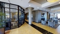 This elegant single-floor space presents a refined and contemporary design, immediately commanding attention with its grand lobby entrance. The entry foyer features large-format polished stone tile flooring in warm gold and deep black tones, anchored by a striking architectural column with a flared cap detail. Sleek glass-panel doors with dark metal framing elegantly separate the entry vestibule from the main interior, while textured stone accent walls and recessed lighting add depth and sophistication throughout.

The elevator bay is framed by richly veined stone cladding and stainless steel doors, contributing to the upscale, professional atmosphere. Adjacent to the elevator area, a seating alcove with lounge chairs invites visitors to pause and relax.

Beyond the entry, the main lounge area opens into a generously proportioned space with soaring ceilings and layered tray ceiling details. Plush grey upholstered chairs are arranged around a central coffee table atop a patterned area rug, creating an inviting seating cluster. Large windows at the far end flood the space with natural light and offer views to an outdoor terrace area. Decorative tall branch arrangements in oversized vessels, curated artwork, and a beverage service station complete the polished and welcoming ambiance of this well-appointed communal space.