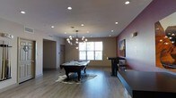 This single-floor recreational space offers a well-appointed and inviting environment designed for social engagement and leisure. The open layout centers around a full-size pool table, positioned atop a striking cowhide-patterned area rug, making it an immediate focal point upon entry. The room is finished with smooth light wood flooring that complements the neutral wall tones, while a bold accent wall painted in a deep wine-red hue adds a layer of visual depth and character. Overhead, a modern globe chandelier serves as a statement lighting fixture, working in harmony with the recessed ceiling lights to ensure the space is well-lit throughout. Natural light filters in through a large window, brightening the interior and providing a connection to the outdoors. Along one wall, a sleek dark-toned bar-height table with matching stools creates an ideal spot for spectators or casual conversation. A vibrant piece of wall art adds a splash of color and personality to the space. A hallway visible in the background suggests additional rooms or amenities beyond, enhancing the overall functionality of this thoughtfully designed communal area.