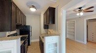 This single-floor residence offers a thoughtfully designed layout that balances classic character with functional living. The galley-style kitchen is a standout feature, lined on both sides with rich dark-stained cabinetry that extends to the ceiling, providing ample storage. Granite countertops run the length of both walls, complemented by a stainless steel sink and black appliances, including a gas range and refrigerator. Distinctive white base cabinets with glass panel doors add a contrasting design detail. The living and bedroom areas are finished with light hardwood flooring and neutral-toned walls, creating a bright and open atmosphere. Crown molding and ceiling fans are present throughout, adding a touch of classic architectural detail. Built-in shelving units provide practical and attractive display or storage space. A generous walk-in closet with rod storage and hardwood flooring offers excellent wardrobe organization. Windows in the living areas invite natural light and outdoor views. The bathroom is finished in clean white tile, featuring a combined tub and shower, a vanity with a mirror medicine cabinet, and a window that provides natural ventilation and light.