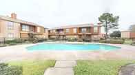 This single-floor residence is part of a wood-paneled, brick-accented multi-building complex that features lush landscaping, a fenced perimeter, and a generously sized communal swimming pool — an inviting outdoor amenity visible from the grounds.

Inside, the interior reflects a thoughtfully curated aesthetic with clean, neutral-toned walls that serve as a backdrop for artistic expression. The dining area is a standout space, anchored by a striking round green marble pedestal table paired with woven rattan chairs atop a natural fiber area rug. A large, framed abstract artwork adorns the wall, adding a sophisticated and gallery-like quality to the room.

The space benefits from recessed ceiling lighting that provides warm, even illumination throughout. Dark hardwood-style flooring runs through the interior, grounding the design with richness and depth. A sliding barn door on a black rail system adds an industrial-chic touch while offering practical room separation. Vintage-style distressed French doors connect interior spaces, contributing character and visual interest.

Overall, the residence balances eclectic design sensibilities with functional layout, creating an atmosphere that is both refined and livable — suited for those who appreciate artful interiors complemented by convenient community amenities.