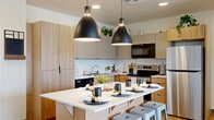 This well-appointed 4-bedroom, 4-bathroom unit is thoughtfully designed to maximize both functionality and comfort across a single floor. The open-concept kitchen and living area serves as the heart of the home, anchored by a central island with a white quartz countertop and seating for multiple residents. Dual matte black pendant lights hang overhead, complementing the warm wood-toned cabinetry, white subway tile backsplash, and stainless steel appliances. Light hardwood flooring runs throughout the main living area, creating a cohesive and inviting atmosphere. The adjacent living room receives abundant natural light through a sliding glass door that opens to a private balcony, offering sweeping views of the surrounding mountains and landscaped grounds. Each of the four bedrooms is carpeted and features a window that brings in natural light, with enough space for a bed and study area — ideal for individual occupancy. Each bedroom is paired with its own dedicated bathroom, finished with modern gray cabinetry, white countertops, and hardwood-style flooring. A hallway connects the private sleeping quarters to the shared living spaces, maintaining a clear and organized layout. This unit provides a practical and modern living experience with quality finishes throughout.