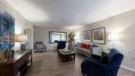 This thoughtfully designed single-floor residence offers a cohesive layout that connects its key living spaces with ease. The expansive living room is a standout feature, showcasing durable wood-look flooring, neutral walls, a textured ceiling, and generous natural light filtering through large windows. A large framed mirror accent adds depth to the room, which comfortably accommodates multiple seating areas.

The galley-style kitchen is well-equipped, featuring gray cabinetry, quartz-style countertops, black appliances including an electric range and dishwasher, and a mosaic tile backsplash. The open sightline from the kitchen through to the living room enhances the sense of connectivity throughout the space.

Adjacent to the kitchen, the dedicated dining area provides an intimate setting beneath a statement pendant light, with a window overlooking what appears to be a balcony or exterior space, allowing natural light to fill the room.

The bedroom is generously sized with plush carpeting, multiple windows dressed in light curtains, a ceiling fan, and the added convenience of two side-by-side reach-in closets offering ample storage.

The bathroom features an extended vanity with quartz countertops, gray cabinetry, a well-lit mirror, and a separate water closet, combining practicality with a clean, modern aesthetic throughout.
