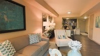 This single-floor residence showcases a thoughtfully designed open-concept layout that connects the living area and kitchen in a seamless, functional flow. The living room is warmly illuminated by recessed lighting and ambient lamps, casting a soft, inviting glow across the space. Neutral-toned walls complement the room's contemporary furnishings, including a plush grey sofa accented with teal patterned cushions and a sleek white accent chair. A decorative console table anchors one wall, styled with elegant table lamps and a geometric mirror that adds depth and visual interest. The open sightline extends into a well-equipped kitchen featuring dark cabinetry, stainless steel appliances, and a dining area, making the space ideal for both relaxed living and entertaining. Light wood-toned flooring runs throughout, unifying the areas and enhancing the sense of continuity. Decorative touches, including sculptural accessories and coordinated soft furnishings, give the space a polished, model-home aesthetic. Overall, the layout is efficiently organized to maximize comfort and style within a compact single-story footprint.