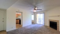 This single-floor unit is thoughtfully arranged to maximize functionality and flow. The layout, as revealed by the 3D floor plan, features a generously sized central living area that serves as the heart of the space, connecting seamlessly to the surrounding rooms. The living room is carpeted and benefits from abundant natural light, with a large window and sliding glass doors that open to an outdoor balcony area, framing views of surrounding greenery. A ceiling fan with an integrated light fixture is centrally mounted on the textured ceiling, enhancing both comfort and ambiance.

Adjacent to the living area, the kitchen is accessible through an open archway, creating a connected feel between the two spaces. The kitchen is equipped with light wood cabinetry, dark countertops, a black range, and a sink positioned beneath upper cabinets, offering a practical and organized workspace. Hard flooring in the kitchen contrasts with the plush carpet of the living area.

The floor plan also indicates additional rooms branching off from the main living space, including what appear to be bedrooms and a bathroom, each with their own window access for natural light. Overall, the unit presents a clean, functional design with a comfortable and livable atmosphere.