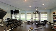 This fitness center offers a comprehensive workout environment designed to support a wide range of exercise routines. The space is appointed with dark rubber flooring, providing both durability and comfort underfoot during training sessions. A full rack of dumbbells lines one wall, offering a broad range of weight options for free weight exercises, while a multi-function cable and strength machine anchors the center of the room, enabling a variety of resistance training movements. An adjustable weight bench adds further versatility to the available equipment. Large mirrors span an entire wall, enhancing the sense of openness and allowing users to monitor their form during workouts. A wall-mounted flat-screen television is positioned for easy viewing from multiple areas of the room. Natural light flows in generously through several windows fitted with horizontal blinds, offering views of the surrounding greenery outside. Recessed lighting in the textured ceiling ensures the space remains well-lit at all hours. The overall ambiance is clean, functional, and purpose-built, making this fitness center a practical and inviting facility for a variety of fitness levels and training goals.