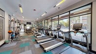 Located at 1015 NW 21st Ave in Gainesville, Florida, this fitness center is a well-equipped, single-floor facility designed to support a comprehensive workout experience. The space is laid out in a long, open-plan format that maximizes usability and flow between equipment zones.

A defining architectural feature is the expansive floor-to-ceiling window wall running along one side of the room, which floods the interior with abundant natural light and offers clear views of the adjacent outdoor pool area. This connection to the outdoors creates an open, energizing atmosphere throughout the space.

The cardio section is prominently featured along the windowed wall and includes a full row of True-brand treadmills, elliptical trainers, and upright stationary bikes — all equipped with digital display consoles. Opposite the cardio zone, the strength training area houses a multi-station cable machine, a Body-Solid functional trainer, a dumbbell rack, and various weight machines, providing a thorough range of resistance training options.

The flooring consists of a patterned carpet tile in a combination of grey and teal tones, lending the space a modern, vibrant aesthetic. Overhead, linear fluorescent lighting ensures the room is consistently and evenly lit. A wall-mounted flat-screen TV is positioned near the entrance for added convenience. Access is controlled via a keypad entry system, and posted fitness center rules are clearly displayed near the entrance.