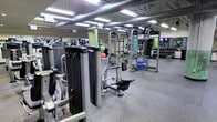 This fitness facility is a comprehensive, single-floor gym designed to accommodate a wide variety of workout needs. The space is expansive and open, with an industrial-style ceiling featuring exposed ductwork and bright fluorescent lighting that ensures excellent visibility throughout the floor.

The strength training area is well-appointed with an extensive selection of selectorized resistance machines, a cable and functional training station, and a full dumbbell rack stocked with a wide range of free weights. Stability balls and other functional training accessories are readily accessible throughout the floor.

The flooring consists of durable rubber matting, providing both safety and comfort underfoot — a practical choice for a high-traffic training environment. Mirrored walls line portions of the space, allowing members to monitor their form during exercises and contributing to an open, airy feel.

The layout is thoughtfully organized to provide clear pathways between equipment stations, promoting a safe and efficient workout flow. Overhead, the exposed industrial ceiling adds a spacious, warehouse-style ambiance that is both modern and functional. Overall, the facility is well-maintained and comprehensively equipped to support strength training, functional fitness, and general conditioning for users of all fitness levels.