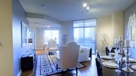 Located in the heart of Dallas, TX, this sophisticated single-floor residence offers a seamlessly open layout that blends the dining and living areas into one cohesive, light-filled space. Expansive floor-to-ceiling windows flood the interior with natural light while framing impressive views of the surrounding city skyline, creating a dynamic urban backdrop that shifts beautifully throughout the day. The living area is anchored by a plush sofa and elegantly styled accent chairs, complemented by refined décor including sculptural botanical arrangements that add an organic touch to the contemporary aesthetic. Rich hardwood flooring runs throughout, adding warmth and continuity to the space. The dining area features a striking high-back upholstered dining set atop a bold geometric area rug, lending a sense of classic elegance with a modern edge. Track lighting installed along the ceiling provides layered illumination, enhancing the open and airy ambiance. The overall design balances sophistication and comfort, making it both a visually impressive and highly functional living space in one of Dallas's vibrant urban settings.