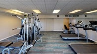 This fitness center offers a comprehensive workout environment spread across a single, open floor. The space is finished with a distinctive patterned carpet in grey and teal tones, providing a clean and modern aesthetic throughout. A drop tile ceiling with recessed panel lighting ensures consistent, even illumination across the room, complemented by generous natural light streaming in through multiple large windows that frame outdoor views of the surrounding landscape.

The cardio zone is well-appointed, featuring multiple treadmills, an elliptical trainer, and a stationary recumbent bike, each positioned to take advantage of the natural light and window views. Wall-mounted monitors are available in this area for entertainment during workouts. A multi-station cable machine occupies a central position in the room, offering a wide variety of strength training exercises for users of all fitness levels. A dedicated free weights section includes a full rack of dumbbells arranged along one wall, accompanied by a workout bench for targeted strength training. The layout is thoughtfully organized to provide ample space between equipment, ensuring safe and comfortable movement throughout the facility. Access to the fitness center is provided through a wood-framed door that connects to an adjacent common area, making it conveniently accessible.