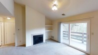 This single-floor space centers around a bright and open living area designed for comfort and functionality. The room is anchored by a tile-surround wood-burning fireplace set against a tall, vaulted wall, lending a sense of architectural height and warmth to the space. Adjacent to the fireplace, built-in open shelving provides convenient display or storage options, adding a structured yet understated design element to the wall.

Soft, neutral-toned carpeting covers the floor throughout the living area, contributing to a calm and inviting atmosphere. The ceiling features a decorative light fixture with an ornate medallion detail, adding a subtle touch of elegance to the otherwise clean and minimal aesthetic.

Natural light fills the room generously through a large sliding glass door fitted with sheer drapery panels. The door opens directly onto a balcony or deck, where mature trees are visible, offering a pleasant and private outdoor view. The overall ambiance is quiet and airy, with warm neutral wall tones and ample natural light creating a welcoming environment throughout the living space.