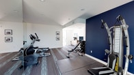 This dedicated fitness space is thoughtfully designed to support a comprehensive workout experience. The room features a clean, modern aesthetic defined by crisp white walls, a bold navy blue accent wall, and durable flooring with protective rubber mats strategically placed beneath the equipment. Recessed lighting illuminates the space evenly, creating a bright and energizing atmosphere. The cardio area is outfitted with multiple machines, including an elliptical and a stationary bike, positioned along one side of the room. A sturdy adjustable weight bench sits at the center, accompanied by a well-stocked dumbbell rack offering a range of free weights for strength training. A cable resistance station mounted on the navy accent wall adds versatility for a variety of functional exercises. Framed artwork adorns the walls, adding a polished touch to the utilitarian space. An open doorway leads to an adjacent area with additional storage and natural light, enhancing the overall sense of connectivity. The layout is efficient and practical, making excellent use of the available floor space while maintaining clear pathways between stations. This single-floor fitness space delivers both form and function in equal measure.