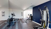 This dedicated fitness space is thoughtfully designed to support a comprehensive workout experience. The room features a clean, modern aesthetic defined by crisp white walls, a bold navy blue accent wall, and durable flooring with protective rubber mats strategically placed beneath the equipment. Recessed lighting illuminates the space evenly, creating a bright and energizing atmosphere. The cardio area is outfitted with multiple machines, including an elliptical and a stationary bike, positioned along one side of the room. A sturdy adjustable weight bench sits at the center, accompanied by a well-stocked dumbbell rack offering a range of free weights for strength training. A cable resistance station mounted on the navy accent wall adds versatility for a variety of functional exercises. Framed artwork adorns the walls, adding a polished touch to the utilitarian space. An open doorway leads to an adjacent area with additional storage and natural light, enhancing the overall sense of connectivity. The layout is efficient and practical, making excellent use of the available floor space while maintaining clear pathways between stations. This single-floor fitness space delivers both form and function in equal measure.