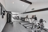 The fitness center at 3900 Fir Tree in Riverside, California, is a well-appointed, single-level facility designed to support a comprehensive workout experience. The space features soaring vaulted ceilings accented by bold dark exposed beams, lending the room a dynamic and modern character. A striking black accent stripe runs along the upper perimeter of the white walls, creating a sharp, contemporary aesthetic throughout.

The main workout area is equipped with multiple True-brand treadmills, elliptical cross-trainers, a stationary recumbent bike, a stair climber, and a multi-function cable machine, providing a thorough range of cardio and strength training options. A dedicated free weight section includes a tiered dumbbell rack stocked with a full set of weights. Full-length mirrored wall panels visually expand the space and allow users to monitor their form during exercise.

Durable rubber flooring covers the entire area, offering both safety and comfort underfoot. Large windows along one side of the room flood the space with abundant natural light while offering direct views of the outdoor pool area, complete with lounge seating, a pergola, and lush landscaping including palm trees. This seamless visual connection to the outdoor amenities creates an energizing and motivating environment for residents.