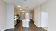 This single-floor residence showcases a thoughtfully designed open-concept layout that seamlessly connects the kitchen and living areas. The kitchen is a standout feature, centered around a white kitchen island with an integrated sink, illuminated by a modern pendant light that adds a contemporary touch. Light wood cabinetry lines the kitchen walls, complemented by stainless steel appliances, including a refrigerator, microwave, and range, providing both style and functionality.

Dark hardwood-style flooring extends throughout the space, creating a warm contrast against the neutral, beige-toned walls and bright white trim. Flush-mount ceiling fixtures provide consistent ambient lighting across the open floor plan, enhancing the airy feel of the interior.

The open corridor beyond the kitchen leads to additional rooms, hinting at a well-organized layout with clearly defined living zones. A white paneled door and a set of closet doors are visible, suggesting practical storage solutions integrated into the design. The overall aesthetic is clean, modern, and move-in ready, with cohesive finishes that lend the space a polished, contemporary character.