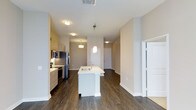 This single-floor residence showcases a thoughtfully designed open-concept layout that seamlessly connects the kitchen and living areas. The kitchen is a standout feature, centered around a white kitchen island with an integrated sink, illuminated by a modern pendant light that adds a contemporary touch. Light wood cabinetry lines the kitchen walls, complemented by stainless steel appliances, including a refrigerator, microwave, and range, providing both style and functionality.

Dark hardwood-style flooring extends throughout the space, creating a warm contrast against the neutral, beige-toned walls and bright white trim. Flush-mount ceiling fixtures provide consistent ambient lighting across the open floor plan, enhancing the airy feel of the interior.

The open corridor beyond the kitchen leads to additional rooms, hinting at a well-organized layout with clearly defined living zones. A white paneled door and a set of closet doors are visible, suggesting practical storage solutions integrated into the design. The overall aesthetic is clean, modern, and move-in ready, with cohesive finishes that lend the space a polished, contemporary character.
