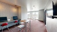 This thoughtfully designed community lounge in Ames, Iowa offers a versatile, single-floor layout that accommodates a variety of social and functional needs. The space features an open lounge area furnished with a neutral sectional sofa, vibrant red accent chairs, and a wooden side table, all anchored by a wall-mounted flat-screen television. Bold accent walls — including a striking red herringbone panel and an industrial-style textured feature wall — add visual character throughout.

A dedicated kitchenette area provides a stainless steel countertop with a sink, wood-toned cabinetry, and a Keurig coffee station set against a large-format stone-look tile backsplash, making it ideal for casual refreshments. Package lockers line one wall near the lounge entrance, offering a practical convenience for residents.

Multiple seating and work areas are distributed throughout the space, including high-top tables with bar stools, a round dining table beneath a statement pendant light, and private nook-style workstations equipped with a desktop computer. Exposed ductwork adds an industrial design touch to select areas.

Large windows flood the interior with natural light and frame views of the surrounding Ames streetscape, including nearby greenery and a waterway. Striped carpet in warm red and neutral tones ties the space together, creating an energetic yet welcoming environment.