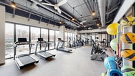 Located at 1325 W Quincy Ave in Englewood, Colorado, this single-floor fitness facility offers a well-equipped, expansive workout environment defined by a contemporary industrial aesthetic. The space features exposed ductwork and structural elements set against a dark ceiling, contrasted by light-colored walls and neutral carpet flooring that lend a clean, open feel throughout.

The cardio zone is positioned along a wall of large floor-to-ceiling windows, providing an abundance of natural light and outdoor views while accommodating multiple treadmills, ellipticals, and stationary bikes, including air resistance cycles. Ceiling fans are distributed throughout this area, enhancing ventilation and comfort during workouts.

The strength training section hosts a variety of cable machines, functional trainers, free weights, and benches. Full-length mirrors line portions of the walls, visually expanding the space and supporting proper form during exercise. A dedicated studio room with carpeted floors, mounted flat-screen displays, and pendant lighting provides a versatile area suitable for group fitness classes or individual training sessions.

Overall, the facility presents a modern, well-lit environment with generous floor space, thoughtful equipment distribution, and an industrial-inspired design that creates a motivating and functional atmosphere for fitness.