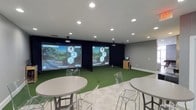 This dedicated indoor golf simulator facility is designed to deliver an immersive and high-performance golf experience in a clean, modern setting. The single-floor space is anchored by two full-size golf simulation bays, each equipped with large projection screens and Foresight Sports launch monitor technology, allowing players to experience true-to-life course play year-round. The artificial turf hitting mats provide an authentic feel underfoot, while ceiling-mounted projectors ensure sharp, vivid visuals during gameplay. Recessed lighting throughout the space creates a bright yet focused atmosphere well-suited for both casual play and serious practice. A small media station with a monitor is conveniently positioned between the bays for easy access to settings or scoring. The lounge area features round high-top tables paired with modern acrylic ghost chairs, offering a stylish and comfortable space for guests to relax, socialize, or watch others play. The neutral gray walls and white trim keep the aesthetic clean and contemporary, while the dark curtain backdrops behind the simulator screens enhance contrast and visual immersion. This facility is well-suited for individual practice sessions, competitive play, group outings, and entertainment events alike.