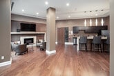 Located at 3 Summerland Terr in Toronto's sought-after M9A neighbourhood, this single-floor amenity space is designed with a refined, contemporary aesthetic throughout. The entrance lobby sets a sophisticated tone with rich hardwood flooring, warm taupe-toned walls, and structural columns finished in a complementary shade. Floor-to-ceiling windows along the perimeter flood the space with abundant natural light while offering views of the lush, landscaped outdoor surroundings. A comfortable seating area near the entrance provides an inviting space for residents to gather or relax. The lounge area serves as a central highlight, featuring a marble-surround fireplace with a wall-mounted television above, surrounded by stylish upholstered armchairs arranged around a round pedestal table — ideal for casual conversation. The full-service kitchen is well-appointed with dark espresso cabinetry, granite countertops, a mosaic tile backsplash, stainless steel appliances, and a double sink. Adjacent to the kitchen, a substantial bar-height dining table with leather seating accommodates group gatherings, illuminated by a row of elegant pendant lights. A large-scale art piece adorns the wall, adding a gallery-like quality to the dining area. The cohesive warm palette and upscale finishes throughout create an inviting, community-focused atmosphere.