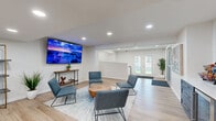 Located at 6006 E 126th St in Grandview, Missouri, this single-floor space offers a thoughtfully designed layout that balances comfort, utility, and modern aesthetics throughout its distinct functional zones.

The main lounge area serves as the centerpiece of the space, featuring light hardwood-style flooring, crisp white walls, and recessed lighting that creates a bright, welcoming atmosphere. A large wall-mounted flat-screen TV anchors the seating area, where a set of contemporary slate-blue accent chairs surrounds a solid wood round coffee table atop a patterned area rug. A sleek console table beneath the TV adds a subtle decorative touch. French doors at the rear of the lounge invite natural light and provide an outdoor connection.

Adjacent to the lounge, a bar-height counter with bold, colorful artwork serves as a casual gathering spot, complemented by black pedestal bar stools and open shelving nearby. A snack and beverage station with navy blue cabinetry and a mini-refrigerator provides convenient refreshments.

The kitchen is clean and functional, outfitted with white shaker cabinetry, a subway tile backsplash, stainless steel appliances including a gas range, microwave, and refrigerator, and a water cooler station.

A glass-door fitness room is accessible from the main corridor, housing cardio and exercise equipment within a dedicated, mirrored workout space.