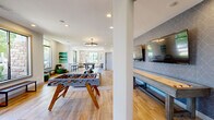 Located at 4505 Dublin Blvd in Colorado Springs, CO, this single-floor community amenity space offers a well-appointed and thoughtfully designed layout spanning a generous footprint. The kitchen area is a standout feature, showcasing full exposed brick walls, white shaker cabinetry, white quartz countertops, open floating shelves, matte black pendant lighting, and an arched window that fills the space with natural light. A central island with bar seating provides a casual gathering spot adjacent to the kitchen.

The open-plan dining and lounge area flows seamlessly from the kitchen, featuring wood-look tile flooring throughout, high ceilings with vaulted sections, and multiple bar-height tables with green upholstered seating. Large windows along the perimeter frame views of the surrounding greenery and bring in abundant daylight.

The living and entertainment area is highlighted by a striking multi-screen media wall set within warm wood slat paneling, complemented by carpeted flooring and comfortable seating. A dedicated game room includes a foosball table, a shuffleboard table, and a pool table, offering diverse recreational options. Throughout, the design balances modern finishes with warm, inviting textures, creating a functional and welcoming community gathering environment.
