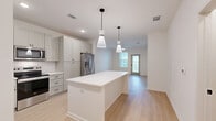 Located at 100 Wilton Cir S in Palm Coast, Florida, this single-floor residence offers a thoughtfully designed layout that maximizes every inch of its compact footprint. The open-concept living area seamlessly connects the kitchen, dining, and living spaces, creating an airy and functional environment ideal for everyday living. Light-toned luxury vinyl plank flooring flows throughout the main living areas, complementing the bright, neutral color palette and recessed lighting that keep the space feeling open and fresh. The kitchen is a standout feature, showcasing gray shaker-style cabinetry with ample storage, a white subway tile backsplash, sleek quartz countertops, and a central island with pendant lighting overhead. Stainless steel appliances, including a French door refrigerator, range, microwave, and dishwasher, complete the well-appointed culinary space. The living and dining area extends toward a glass door that invites natural light and outdoor views. The bedroom is a comfortable retreat featuring plush carpet, a ceiling fan, and windows that bring in natural light, with direct access to the bathroom and closet. Two closets and a dedicated laundry area add practical storage and convenience throughout the home.