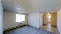 Located at 90 South 18th Avenue in Brighton, Colorado, this single-floor unit offers a straightforward and functional layout well-suited for everyday living. The living area is finished with wall-to-wall carpet in a neutral grey tone, providing a comfortable and cohesive foundation throughout the space. A large window allows natural light to fill the room, creating a bright and open atmosphere. The walls are painted in a clean, neutral white, contributing to a simple and versatile interior palette. A textured popcorn ceiling adds a classic character to the space. Climate control is provided by a wall-mounted air conditioning unit and a baseboard heating system, ensuring year-round comfort. The living area transitions smoothly into an adjoining hallway that connects to additional rooms, suggesting a compact yet well-organized floor plan. The hallway features tile flooring, offering a durable and easy-to-maintain surface in the transitional spaces. Overall, the unit presents a clean, no-frills interior that offers practical functionality in a residential setting within the Brighton community.