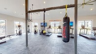 This fitness center is a comprehensively outfitted facility designed to accommodate diverse workout preferences. The main exercise area features soaring vaulted ceilings with exposed beam accents and recessed lighting, creating a bright, open atmosphere. Large windows and glass doors along the perimeter flood the space with natural light while offering outdoor views. Gray tile flooring throughout provides a durable, clean foundation.

The cardio zone is lined with multiple treadmills and elliptical machines, each equipped with individual monitors for entertainment. A dedicated functional training rig supports TRX suspension systems and an Everlast heavy punching bag, while motivational "Hustle & Muscle" wall graphics add energy to the environment. The strength training area includes adjustable benches, a full rack of dumbbells and kettlebells, a wall-length mirror, and additional motivational wall decals. A ceiling fan ensures comfortable airflow throughout the space.

A separate yoga and barre studio features warm hardwood flooring, floor-to-ceiling mirrors with mounted barre rails, and large windows with outdoor views. Inspirational Rumi quotes adorn the walls, promoting mindfulness and calm. The facility also includes accessible, gender-separated restrooms and a dual-station water fountain with a bottle-filling station for added convenience.