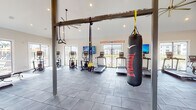 This fitness center is a comprehensively outfitted facility designed to accommodate diverse workout preferences. The main exercise area features soaring vaulted ceilings with exposed beam accents and recessed lighting, creating a bright, open atmosphere. Large windows and glass doors along the perimeter flood the space with natural light while offering outdoor views. Gray tile flooring throughout provides a durable, clean foundation.

The cardio zone is lined with multiple treadmills and elliptical machines, each equipped with individual monitors for entertainment. A dedicated functional training rig supports TRX suspension systems and an Everlast heavy punching bag, while motivational "Hustle & Muscle" wall graphics add energy to the environment. The strength training area includes adjustable benches, a full rack of dumbbells and kettlebells, a wall-length mirror, and additional motivational wall decals. A ceiling fan ensures comfortable airflow throughout the space.

A separate yoga and barre studio features warm hardwood flooring, floor-to-ceiling mirrors with mounted barre rails, and large windows with outdoor views. Inspirational Rumi quotes adorn the walls, promoting mindfulness and calm. The facility also includes accessible, gender-separated restrooms and a dual-station water fountain with a bottle-filling station for added convenience.