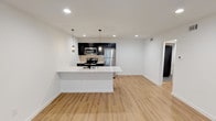 This thoughtfully designed single-floor unit showcases a clean, modern aesthetic carried consistently throughout every room. Light-toned hardwood-style flooring runs continuously across the open-concept living and kitchen area, creating a bright and cohesive feel that is further enhanced by crisp white walls and recessed lighting overhead.

The kitchen is a standout feature, anchored by a generous peninsula with a white quartz countertop, an undermount sink with a matte black faucet, and seating space along one side. Dark cabinetry provides a bold contrast against the light surroundings, while stainless steel appliances — including a refrigerator, range, and built-in microwave — add a polished, functional touch. A textured tile backsplash and elegant pendant lighting complete the space. The open layout flows seamlessly into a generous living area, which benefits from natural light through a window fitted with blinds.

The bedroom offers a quiet, well-proportioned retreat with its own window and direct access to additional spaces via striking matte black interior doors. One of these doors leads to a dedicated laundry closet with a stacked washer and dryer unit.

The bathroom is finished to a high standard, featuring large-format white wall tiles, a glass-enclosed walk-in shower with a dark penny-tile floor, and matte black hardware accents that tie the design together cohesively.