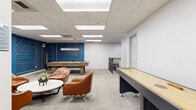 Located at 1101 Iroquois Ave in Naperville, Illinois, this single-floor amenity space is thoughtfully designed to serve as a social and recreational hub. The main lounge area is anchored by plush cognac leather lounge chairs arranged around a round white coffee table, creating an inviting gathering spot. A high-top wood table with black bar stools adds a casual dining or social element nearby. The space transitions seamlessly into a dedicated game room, where a full-size pool table and a regulation shuffleboard table take center stage against a striking blue accent wall with geometric wood detailing — a bold focal point that adds character and visual depth. Overhead, a drop tile ceiling with recessed and panel lighting ensures the space is well-lit throughout. Neutral, multi-toned carpet tiles in the lounge and game areas complement the clean white walls and modern aesthetic. The adjacent kitchen is fully outfitted with warm cherry wood cabinetry, dark countertops, stainless steel appliances including a refrigerator, dishwasher, range, and microwave, and a built-in breakfast bar with seating. Large porcelain floor tiles complete the kitchen's polished, functional design. Together, these areas create a versatile and well-appointed space suited for both relaxation and entertainment.