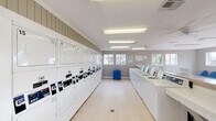 This community laundry facility on South Padre Island Drive in Corpus Christi, Texas, offers a clean, functional, and well-organized space designed to serve residents efficiently. The single-floor layout is bright and open, illuminated by overhead fluorescent lighting and supplemented by natural light streaming through multiple white-framed windows positioned along the walls.

The main laundry area features a generous row of stacked commercial dryers lining one wall, numbered for easy identification, alongside a full lineup of top-load washing machines on the opposite side. Each machine is equipped with digital controls for convenient operation. A folding counter provides a practical surface for managing laundry, and a utility bin is centrally placed for easy access.

A dedicated waiting area offers rows of blue molded chairs positioned beneath large windows that overlook the surrounding outdoor grounds, providing a pleasant view while residents wait. The gray vertical wood-panel walls give the space a cohesive, casual aesthetic, complemented by light-toned tile flooring throughout. Crown molding adds a subtle finishing touch to the interior.

A white paneled door at one end of the facility opens to the exterior, where views of a fenced pool area and surrounding landscaping are visible. Ceiling fans help maintain airflow and comfort throughout the space. Service information postings are displayed on the walls for resident reference.