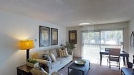 This single-story space centers around a welcoming living room that blends comfort with a casual, well-organized aesthetic. The room features wall-to-wall carpeting in a neutral tone, contributing to a warm and cozy atmosphere. A textured popcorn ceiling adds a classic architectural detail overhead, while the walls are kept in a clean, light neutral hue that enhances the sense of openness.

Natural light flows generously into the space through a full-length sliding glass door fitted with vertical blinds, offering a view of a private outdoor area with greenery visible beyond. This connection to the exterior brightens the room considerably and creates an inviting indoor-outdoor flow.

The living area is furnished with a light-toned sofa accented by decorative throw pillows, paired with a dark rectangular coffee table. A floor lamp in the corner provides warm supplemental lighting. Three framed vintage-style map prints are displayed on the walls, adding a distinctive decorative focal point. A potted plant near the sliding door introduces a natural element to the interior. Overall, the space presents a tidy, functional layout suited for comfortable everyday living.