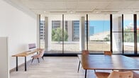 This contemporary single-floor space offers a bright, open-plan layout designed for collaboration and productivity. The defining architectural feature is the expansive wall of floor-to-ceiling windows along one side, framed by sleek concrete columns and fitted with slim black mullions. These large-scale glazed panels flood the interior with abundant natural light while providing sweeping views of the surrounding urban skyline, adjacent green courtyard, and architectural landmarks beyond.

The interior is finished with light-toned wood plank flooring that complements the clean white walls and a recessed panel ceiling fitted with evenly spaced downlights. Multiple rectangular wooden tables are arranged throughout the space, each accompanied by plush tufted velvet chairs in blush pink and warm cognac tones, lending a refined yet approachable aesthetic.

Decorative framed art prints adorn the interior walls, adding a curated touch to the otherwise minimal design. A set of double doors with metal push bars provides access at the rear of the space, while a secondary workstation setup is positioned along the inner wall. The overall ambiance is professional yet inviting, combining modern design sensibilities with generous natural light, urban views, and flexible seating arrangements suitable for a variety of uses.