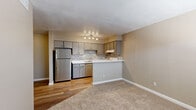 This single-floor residence offers a practical and well-designed layout centered around an open-concept kitchen and living area. The kitchen is equipped with gray cabinetry, a mosaic tile backsplash, light countertops, a stainless steel refrigerator, and a white electric range. A peninsula-style breakfast bar separates the kitchen from the adjoining living space, creating a natural flow between the two areas. Track lighting overhead provides focused illumination throughout the kitchen. The living area features warm beige carpeting that contrasts with the wood-look flooring in the kitchen, giving each zone a distinct character while maintaining a cohesive neutral color palette throughout. The bedrooms are carpeted and finished in soft, warm tones, with each room offering window access for natural light. At least one bedroom includes a walk-in closet with a built-in shelving and hanging rod system, providing practical storage. The bathroom features a full-size soaking tub with a floor-to-ceiling tiled surround accented by a decorative mosaic tile border, complemented by wood-look flooring and a vanity with light countertops. The overall aesthetic is clean, neutral, and move-in ready, with white trim and baseboards providing crisp definition throughout each room.